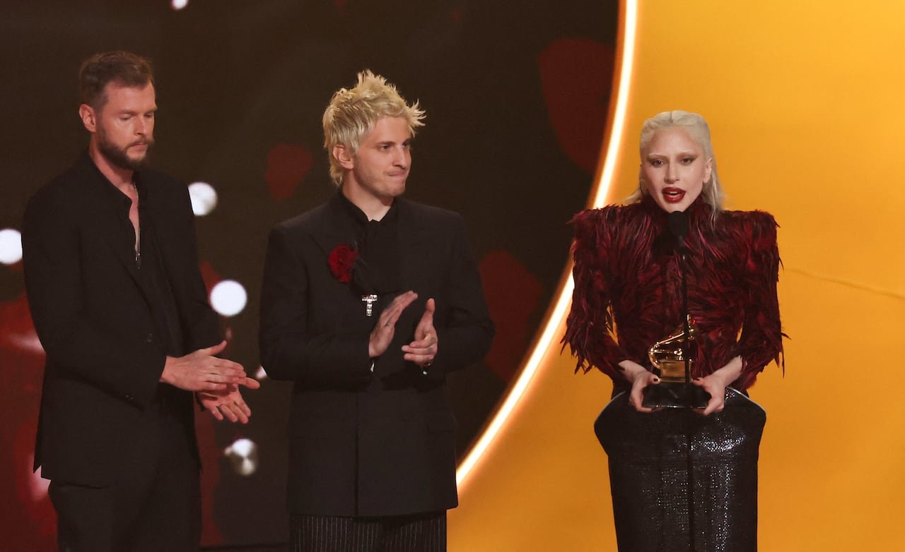 Two men in dark suits standing next to a woman with slicked-back blonde hair in a red and black avant-garde outfit, holding a Grammy award onstage.