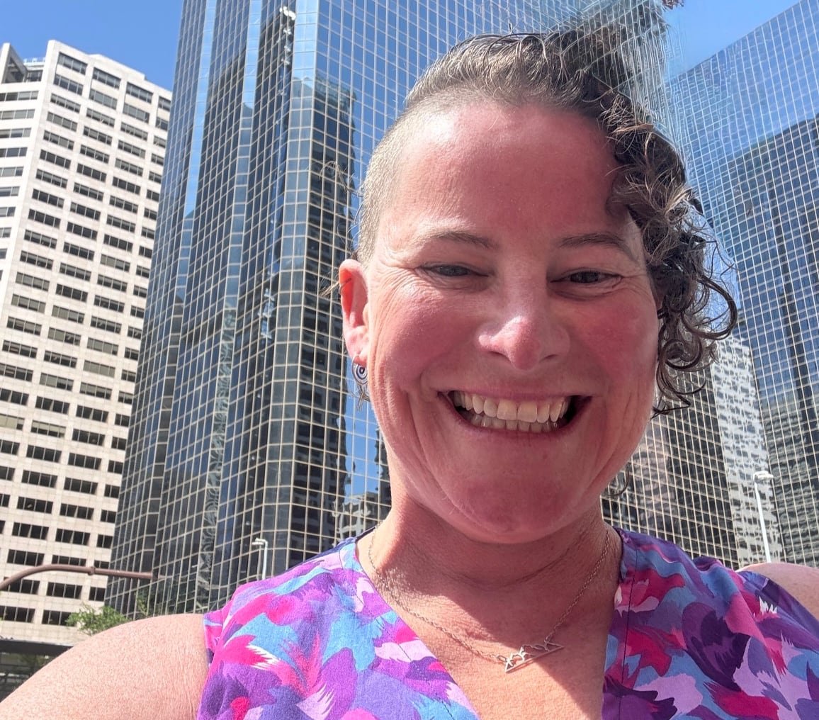 A woman with short curly hair shaved on one side smiles broadly with tall office buildings in the background.