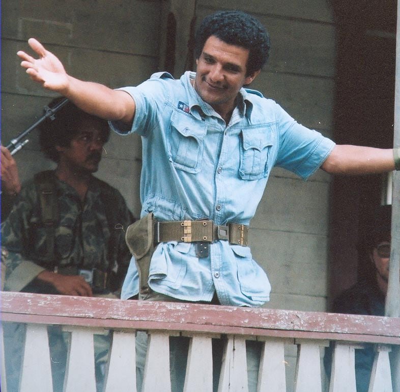 A man in baby blue military fatigues stands on a balcony, smiling and holding both hands out