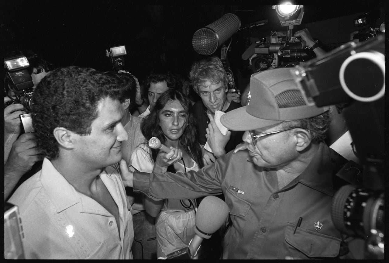Black and white photo of a smiling, curly haired man talking to a bespectacled man in military fatigues, surrounded by reporters with microphones and cameras